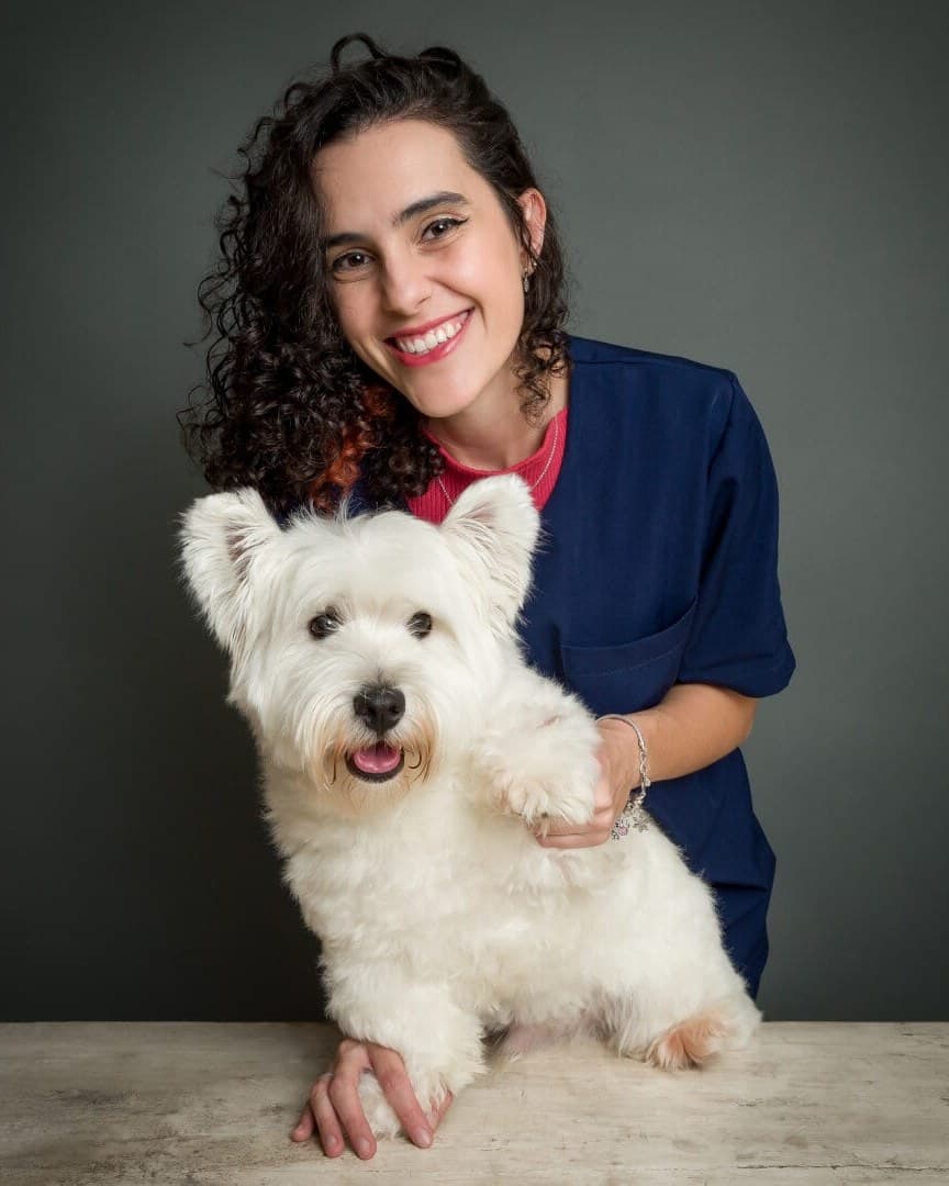 A Dra. Anna Luiza Pollon sorrindo, de jaleco azul, com uma westie branca apoiada na mesa.